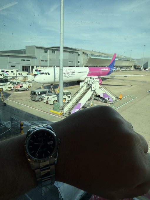 A person wearing a Rolex watch poses in front of an airport window, with a Wizz Air airplane visible on the tarmac, capturing the "see you on the other side" sentiment of an upcoming flight or farewell at the gate.