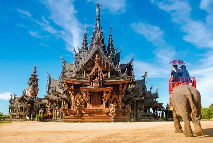 An ornate wooden temple, resembling the Sanctuary of Truth in Pattaya, Thailand, stands against a bright blue sky, with two tourists riding an elephant in the foreground.