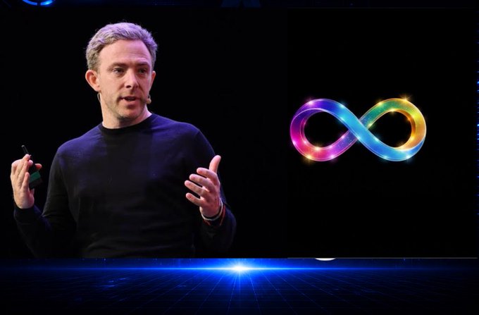 A man giving a presentation on a stage with a dark background, gesturing while speaking next to a brightly colored, glowing rainbow infinity symbol.