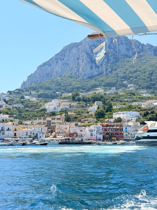 A scenic view from a boat approaching a coastal Mediterranean town with white buildings nestled at the base of a large rocky mountain under a blue sky.