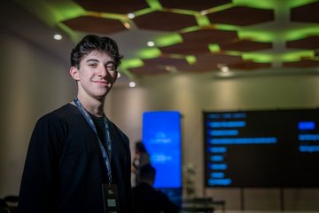 A young man with dark hair and a black sweater smiles while standing in a room with a distinctive honeycomb-patterned ceiling and screens displaying blue charts in the background.