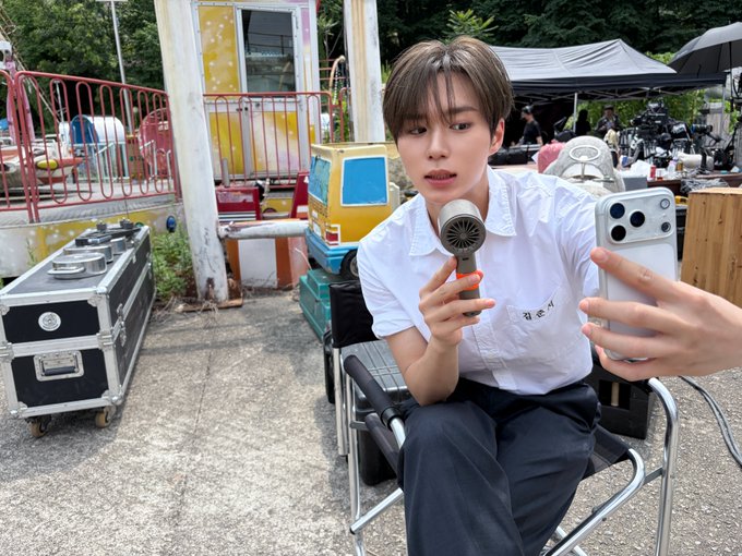 A young man in a white shirt holds a microphone while filming himself with a smartphone, seated in an outdoor filming setup with equipment cases and tents in the background. The Korean text "잡초아" (meaning "weed") is visible on his shirt, aligning with the post's hashtags referencing his content series.