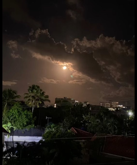 A nighttime photograph of a bright full moon partially hidden behind clouds, with palm trees and a town skyline visible in the foreground.