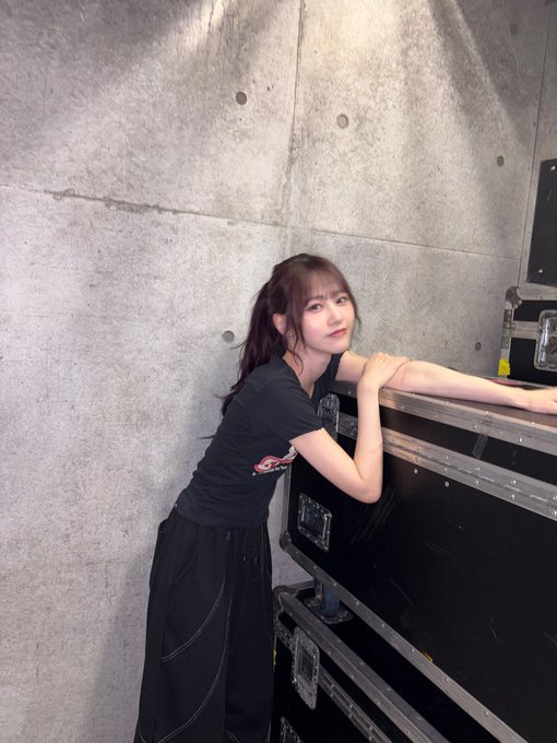 A young woman with long dark hair poses casually in a black t-shirt and skirt, leaning against stacked black equipment cases in what appears to be a backstage or industrial setting, complementing the "ALT" text suggesting an alternative style or music scene context.