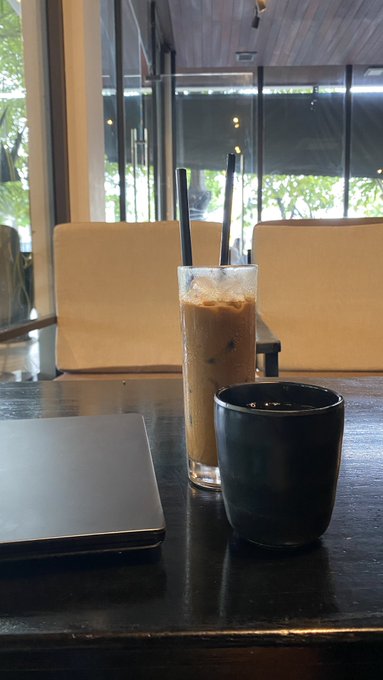 A tall glass of iced coffee with two black straws and a black ceramic mug sitting on a dark wooden table, with a laptop and light-colored chairs visible in the background.