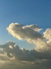 A bright sun shines through fluffy white clouds against a deep blue sky.