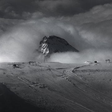 A dramatic black-and-white landscape photograph showing a snow-covered mountain peak emerging from thick clouds and mist, with ski lift structures visible on the snowy slopes below.