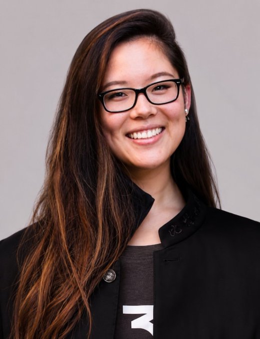 A smiling woman with long dark hair and black-rimmed glasses, wearing a dark blazer over a graphic t-shirt, poses for a professional headshot against a neutral gray background. The portrait appears to accompany a post for "Samson Zhao," likely showcasing her as a team member, professional, or featured individual.