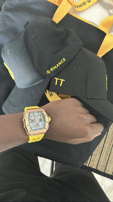 A wrist wearing a yellow Richard Mille-style watch with a Binance cap and 'TT' cap visible in the background.