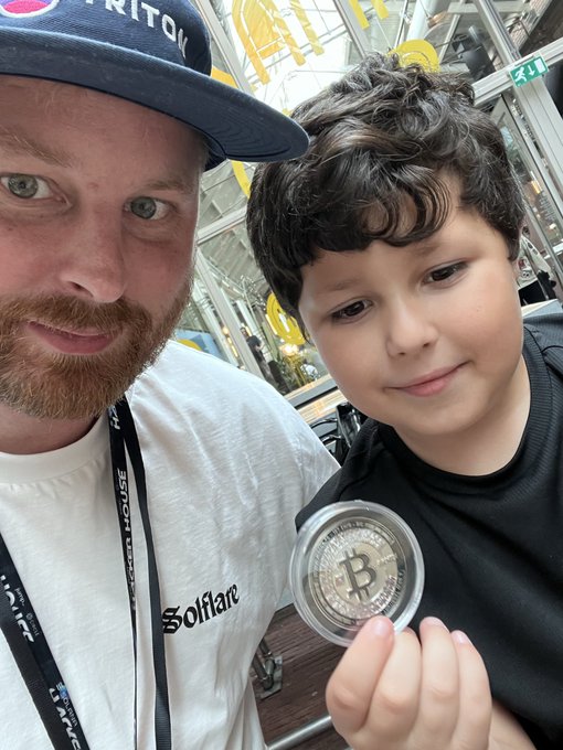 A man wearing a 'Bitcoin' cap and a white 'Solflare' t-shirt poses for a selfie with a young boy who is proudly holding up a physical Bitcoin coin.