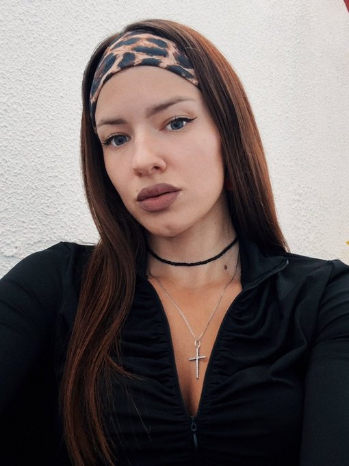 The image features a woman with long brown hair wearing a black top, sitting against a white wall. She has a serious expression as she looks directly at the camera. The photo also includes a pendant necklace around her neck, which adds an elegant touch to her appearance.