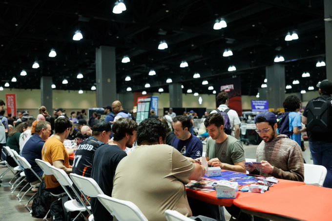 A crowd of people gathered at long tables in a large convention hall, appearing to play trading card games or board games together under bright overhead lighting.