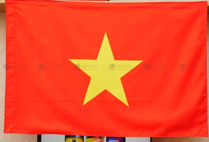 A Vietnamese flag with a large yellow star centered on a red background, featuring 'HẢI TRIỀU' watermarks across the image.