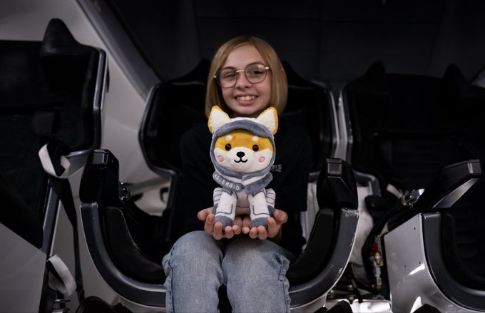 A young girl wearing glasses sits in a space capsule seat, proudly holding a plush Shiba Inu astronaut toy dressed in a hoodie and space suit, embodying her creation of the character. The image visually represents Liv Perrotto, the 11-year-old designer behind the $Asteroid Shiba astronaut, showcasing the real creator with her inspiring space-themed mascot.