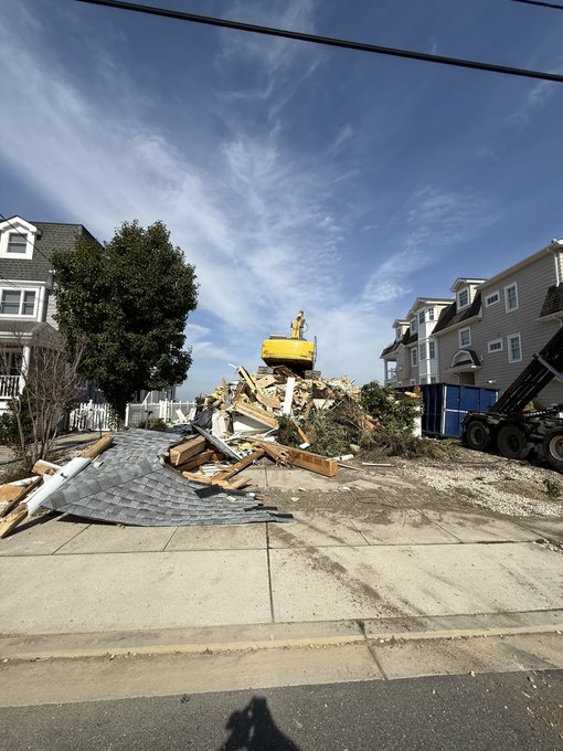 A residential street scene showing a demolished house with a yellow excavator working on a pile of debris, with intact houses on either side under a blue sky.
