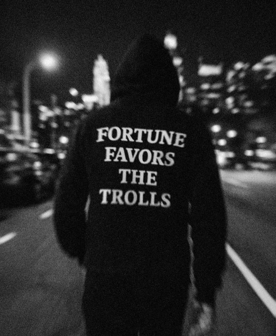The image shows a person walking down a street at night, wearing a black hoodie with white text that says "F Fortune favors the Trolls". The person appears to be carrying a backpack as they walk along the road. The photo has a vintage feel due to its black and white color scheme, which gives it an artistic and nostalgic appearance.
