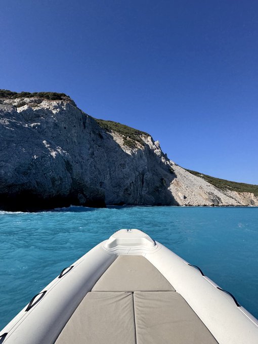 A view from the bow of a white inflatable boat looking toward a dramatic white limestone cliff rising from bright turquoise sea water under a clear blue sky.