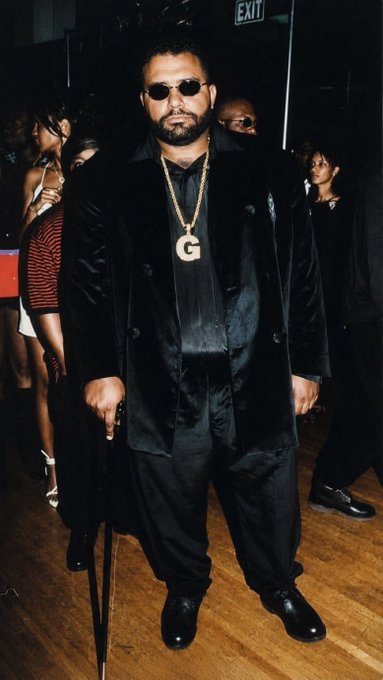 A man wearing a black suit, sunglasses, and a large gold 'G' pendant stands at an event with an 'EXIT' sign visible behind him.