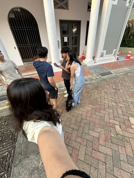 A group selfie taken outside a building with white columns, showing a hand in the foreground and four other people gathered together holding what appears to be a controller.