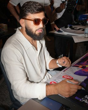 A bearded man wearing sunglasses and a white shirt signs a poster with a black marker, with the text "Crypto.com" visible on the document. The scene appears to be a promotional event, aligning with the post about adding an Arman UFC signature to CryptoCom UFC331 posters.