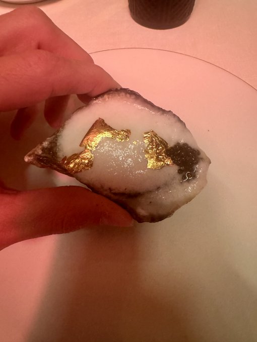 A hand holds a translucent stone or resin slice with irregular gold-colored flakes embedded inside over a warm pink surface.