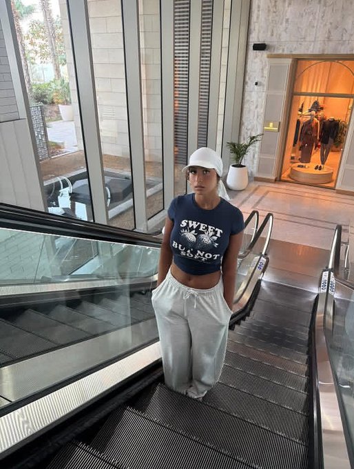The image features a woman standing at the bottom of an escalator inside a building. She is wearing a white hat, a blue shirt with "Sweet" written on it, and white pants. The surrounding area includes a glass wall that allows natural light into the space. There are also potted plants visible in the scene.

The image is related to the post because it contains text about an event called "Put Your Region on the Map with Opinion! The opportunity is here for community builders, sports enthusiasts, and growth leaders to make an impact with @opinionlabsxyz". This event seems to be taking place in a building that has escalators.