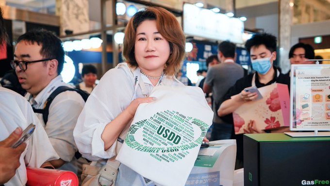 An Asian woman in a white shirt posing at what appears to be a trade show or expo, holding a promotional bag displaying the USDT (Tether) logo made from various smaller cryptocurrency-related words.