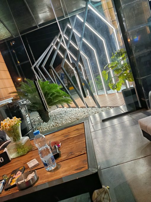 An interior view of a stylish venue featuring a wooden table with flowers, a water bottle, and decor, looking out through a geometric LED-lit archway toward decorative plants and pebbles.