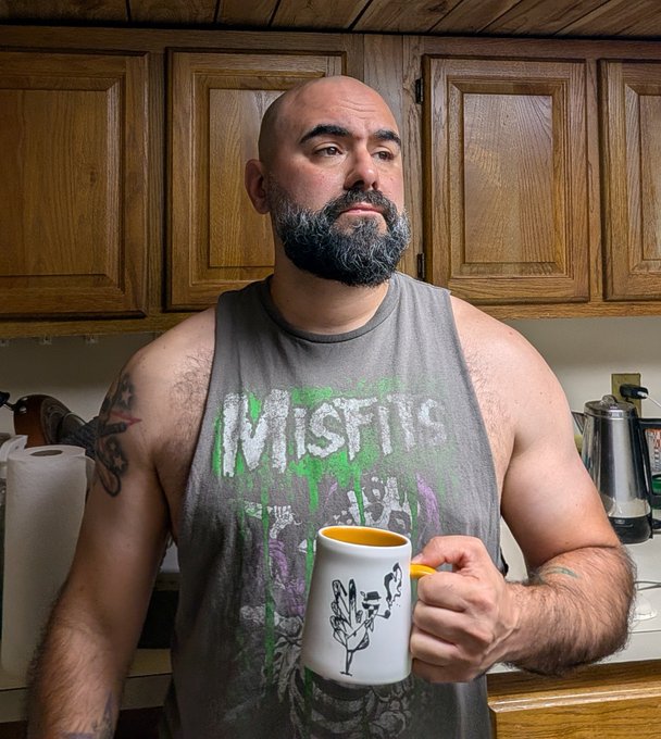 A bearded man wearing a gray Misfits tank top stands in a kitchen holding a coffee mug, posing thoughtfully to match the "GM deep thoughts" caption about reminiscing over an NFT collection like an old yearbook.
