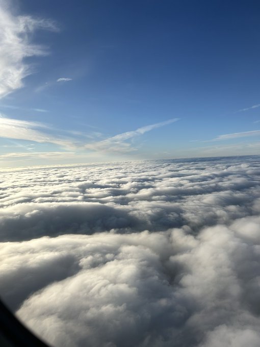 An aerial photograph of a thick cloud layer viewed from an airplane window under a clear blue sky.
