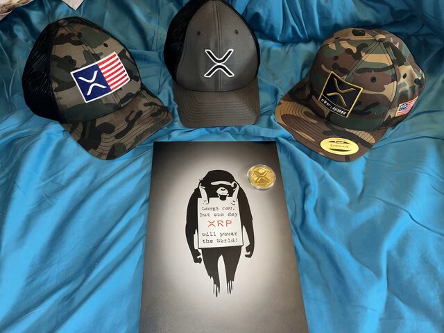 The image features a blue bedspread with three hats placed on top of it, including a camo hat, an American flag hat, and a Batman hat. In front of these hats are two books - one titled "I Love You" by XRP founder and CEO, and another titled "The Art of War". The text in the image reads: "Happy Birthday XRP!!!", which is likely related to the hats or the books on display.