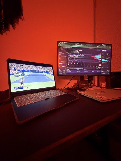 A desk setup with a laptop showing a tennis match and a larger monitor displaying a stock trading chart, illuminated by red ambient lighting.