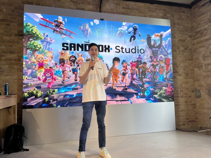 The image features a man standing in front of a large screen displaying a video game called Sandbox Studio. The man appears to be giving a speech or presentation about the game at an event, possibly a conference or gathering related to gaming. He is wearing a white shirt and blue jeans, and he holds a microphone as if addressing an audience.