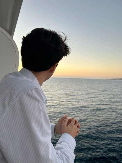 A man in a white striped shirt looks out over the ocean at sunset from what appears to be a boat or ship window, his hands clasped together.