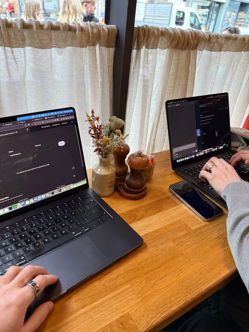 Two people working on laptops at a wooden table in a cafe, with a pepper grinder and small vase of dried flowers between the devices.