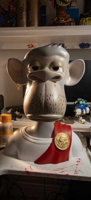 The image features a white bust of a monkey with a red ribbon around its neck, sitting on top of a desk or table. The monkey's face has a sad expression as if it is bored out of its mind. In front of the monkey bust, there are two bottles and a computer monitor. A post on social media reads "Gm gm friends happy hump day! Today we are working on the 22ct fine gold leaf on the 50cm @BoredApeYC bust!". The image seems to be related to an art project or a creative display of monkey figurines, with the sad expression of the monkey as part of the overall theme.