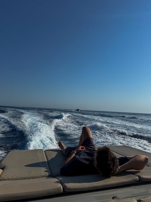 A young man lies on a cushioned surface at the back of a boat, looking out at the ocean wake under a clear blue sky.