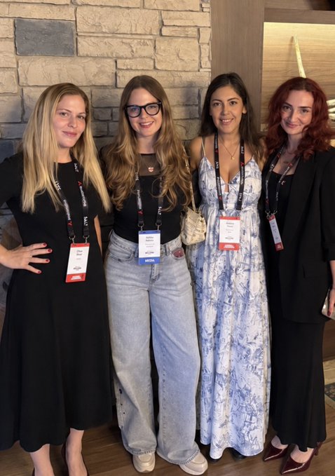 Four women wearing lanyards with name badges pose together in front of a stone wall at what appears to be a conference or event.