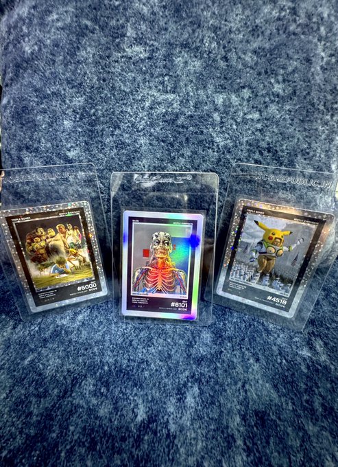 Three Beeple "Everydays" NFT cards (#5000, #6101, #4518) are displayed in clear protective sleeves on a textured blue surface. The holographic cards feature iconic Beeple artwork, including a skeletal figure and the famous Pikachu piece, capturing the digital artist's renowned crypto-art collection.