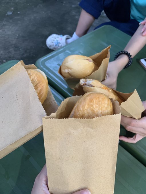 A close-up photo of paper bags filled with bread rolls and small sandwiches, held outdoors near a green table where a child in Crocs is seated.