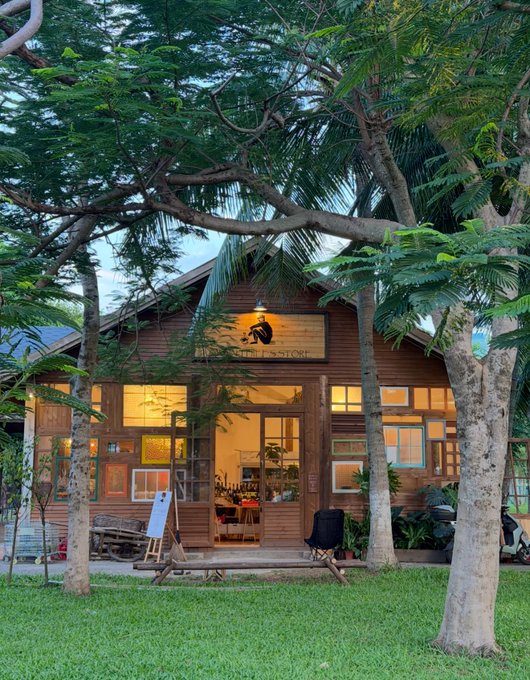 A rustic wooden coffee shop with a sign reading "PHU LONH CAFE" is nestled among lush green trees, its warmly lit windows glowing at dusk.