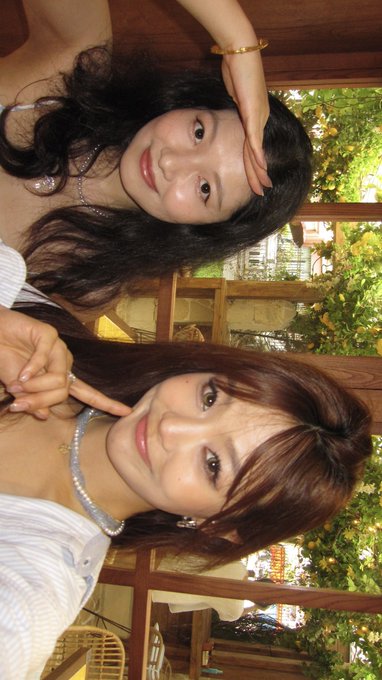 Two women taking a selfie together, both making peace signs and posing with their fingers near their faces in an outdoor setting with greenery.