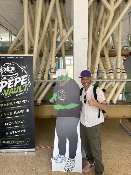 In the image, a man wearing a white shirt and green pants stands next to a cardboard cutout of a person dressed as a zombie with a purple hat. The cardboard cutout appears to be made from paper or cardboard, possibly for an event or promotional activity related to the Rare Pepes store in the background. 

The post features a cartoon image of a man giving a thumbs up and has text that reads "pass by the Rare Pepes... keep building @DJPEPE_ @ScrillaVentura". The cardboard cutout, along with the man's pose, seems to be part of an event or promotional activity related to the store.