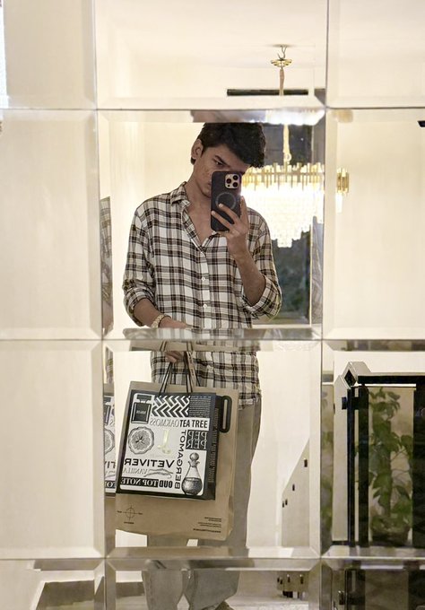 A young man takes a mirror selfie in a tiled, upscale interior with a chandelier behind him, holding a tote bag printed with the text "GOOD MORNING TEA TREE," "OKAY I'M READY," and "OILY SKIN CARE," fitting the cheerful "Good morning everyone" greeting with a stylish, well-groomed look.
