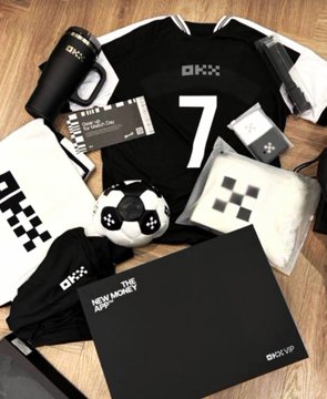 An OKX VIP merchandise bundle laid out on wood, featuring a black jersey with number 7, a soccer ball, socks, and a 'NEW HOME APP' booklet.