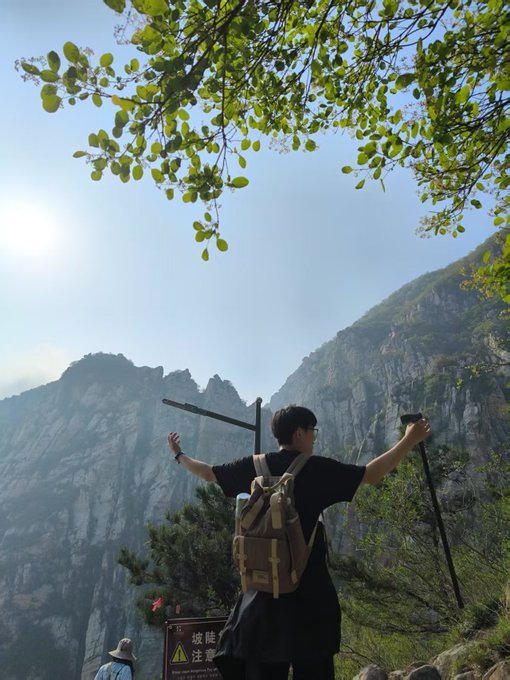 A person wearing a backpack stands with arms outstretched holding a trekking pole, facing a majestic mountain landscape with a tree branch overhead and a warning sign in the foreground reading '坡陡 注意' (Steep slope, caution).