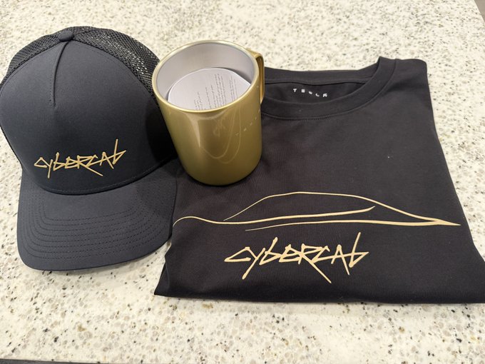 Black "CYBERTRUCK" branded apparel laid out on a granite countertop, including a black cap with gold logo, a gold mug, and a black t-shirt with the Cybertruck outline and logo.