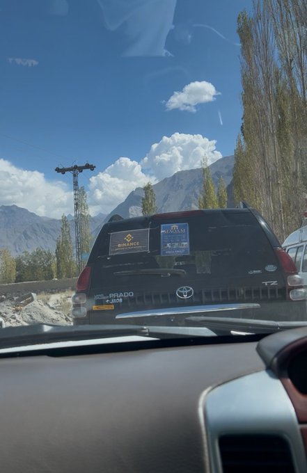 View from inside a car looking at a black SUV with a 'BINANCE' sticker on the rear window, set against a mountainous landscape with tall poplar trees.