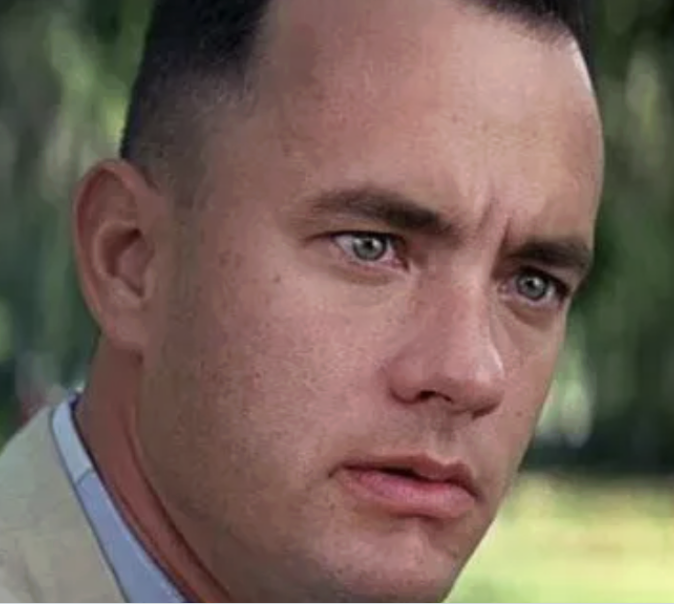 A close-up still of actor Tom Hanks (as Forrest Gump) with a serious expression, set against a blurred green outdoor background.