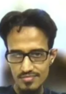 A close-up portrait of a young man with dark hair, a mustache and goatee, wearing glasses and looking at the camera.